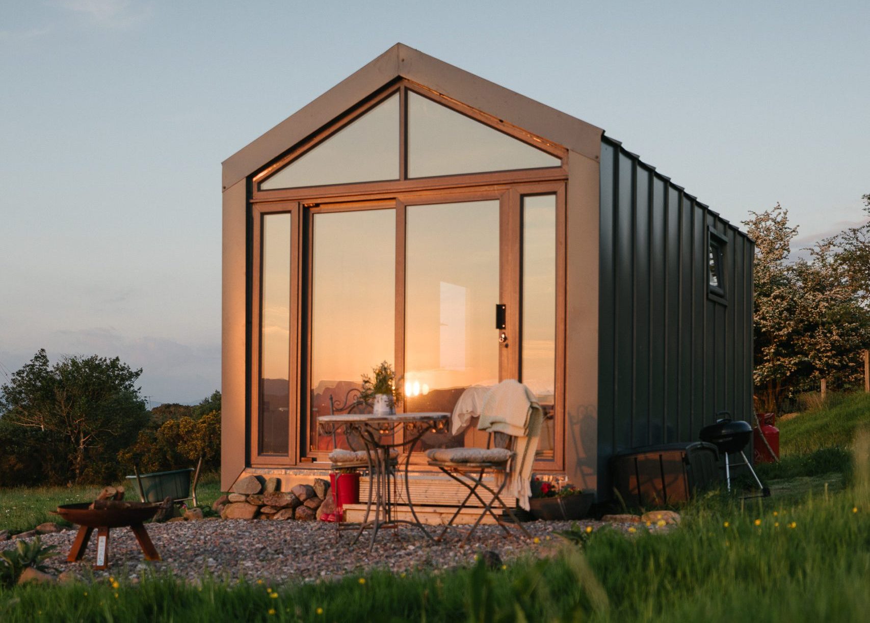 Cabin with a pitched glass gable lit by low evening sun and a fire pit outside