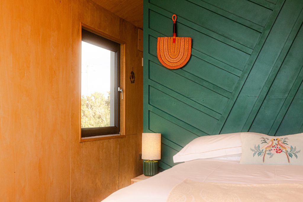 Bed against a deep green panelled wall with a woven fan and bedside lamp